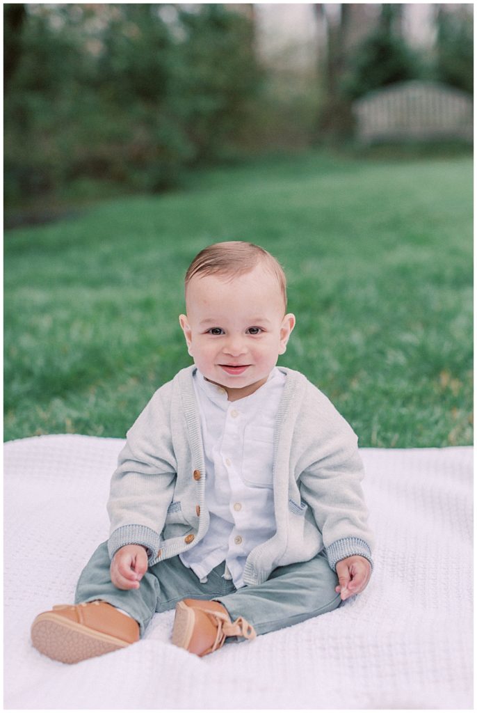 Young baby boy sits on a blanket in a park.