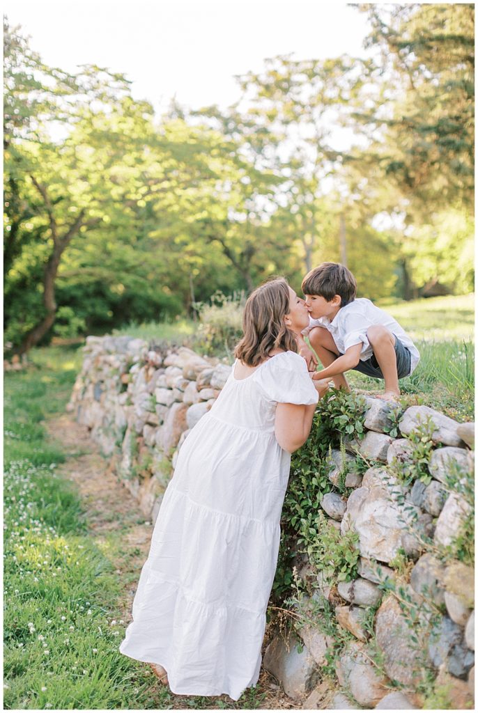 Mother leans over a stone fence to kiss her son during their family photo session