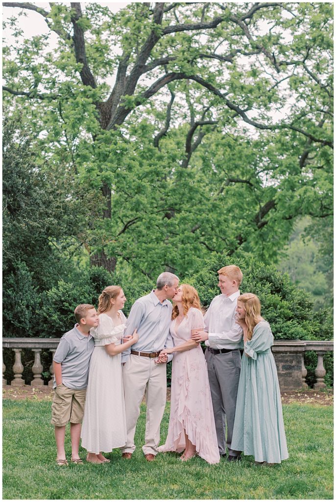 Mother and father kiss while standing with their grown children at Glenview Mansion in Maryland