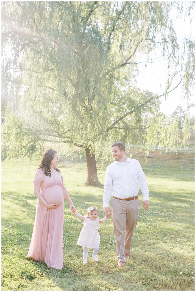 Mother and father walk with their toddler daughter in a field during their Maryland maternity session