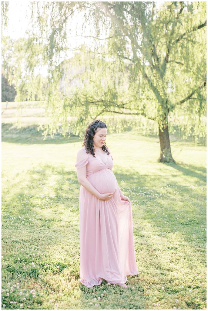 Maternity Photographer Maryland | Pregnant woman stands in a field holding her pink dress
