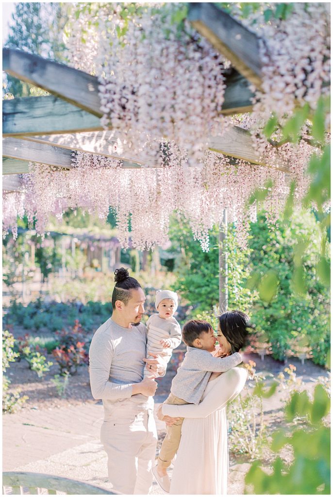 Family cuddles together under purple wisteria at Brookside Gardens during their family photo session