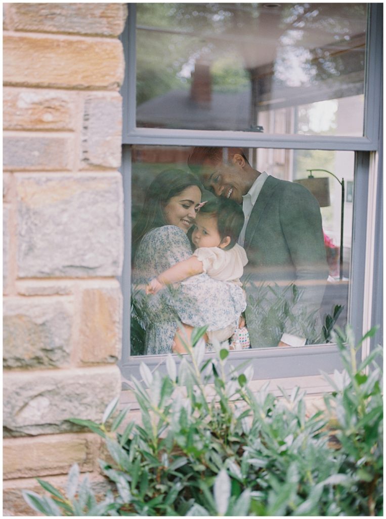 Family stands in their window and snuggles their one year old daughter