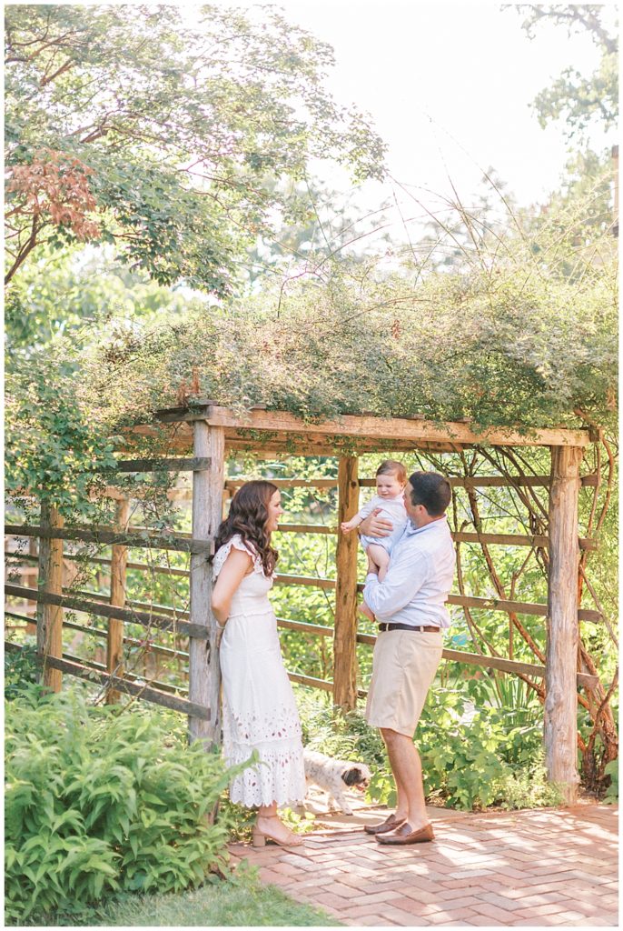 Mom leans up against pergola while dad holds baby boy at Tudor Place