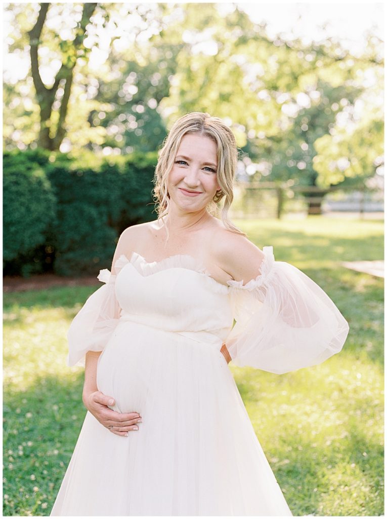 Pregnant woman holds her belly with one hand and smiles at the camera