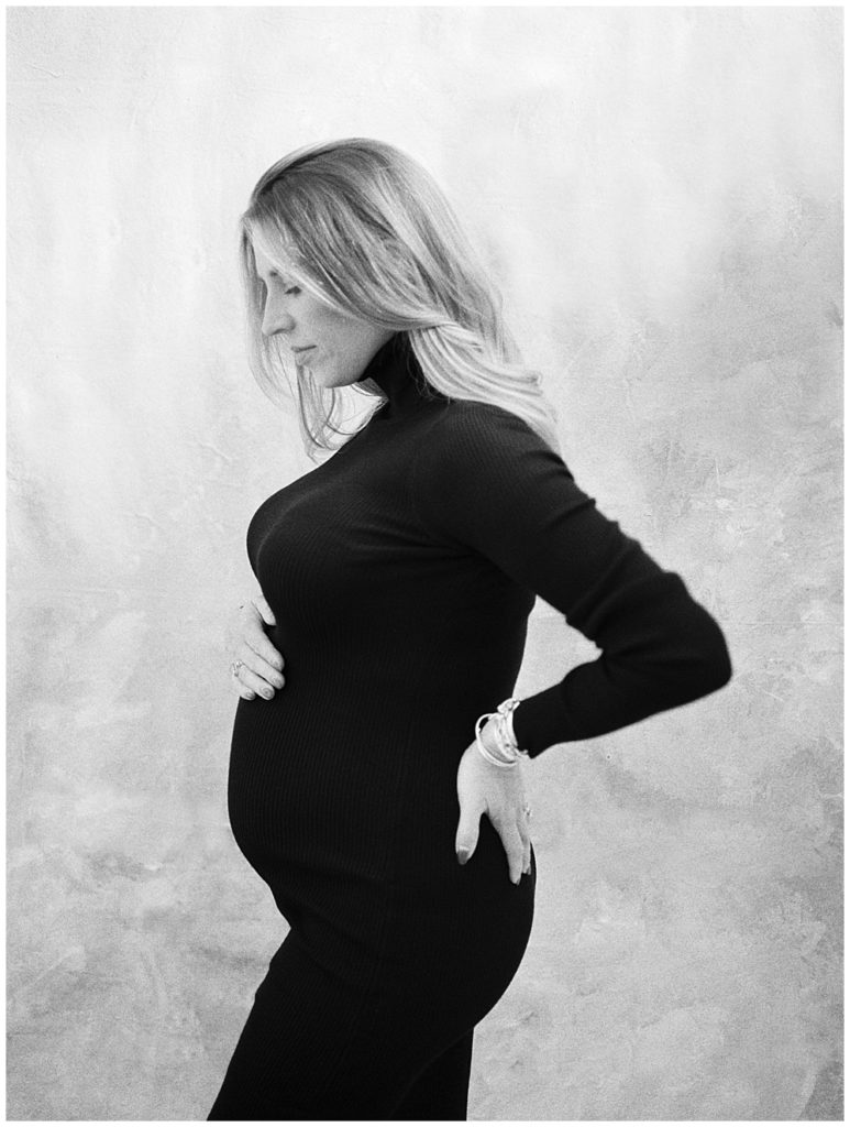 Pregnant mother stands with one hand below belly and one hand behind back during her studio maternity session