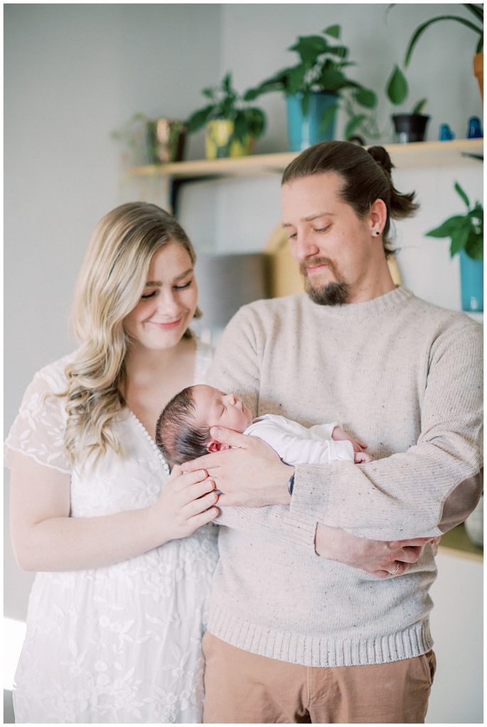 A father holds his newborn daughter while the mother stands next to them with one hand on her husband's hand looking at their baby