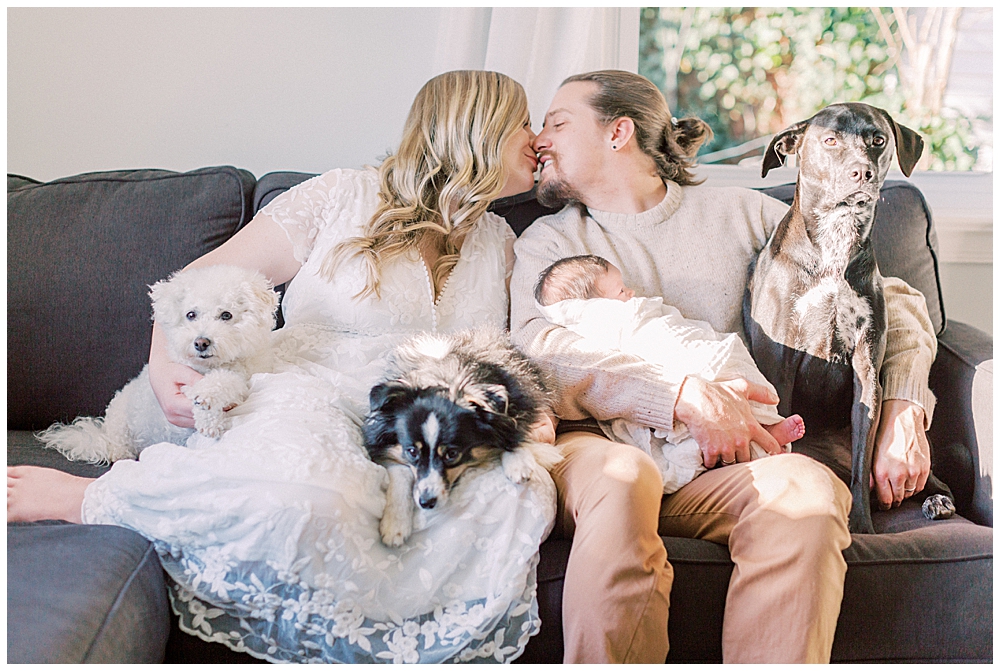 Mother and father sit on their couch with their newborn baby and three dogs and kiss one another