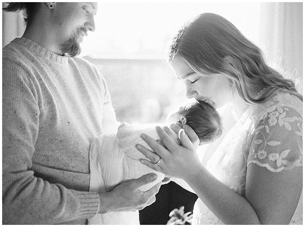 A father holds his newborn daughter in front of him while the mother leans over to kiss the newborn's head