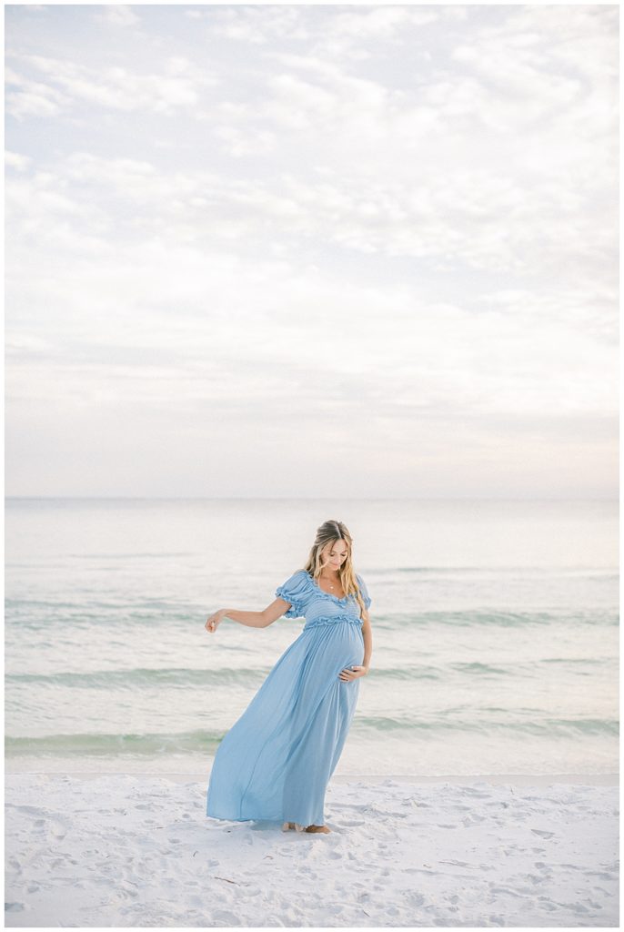 Pregnant mother stands on the beach in a blue dress that is blowing in the wind