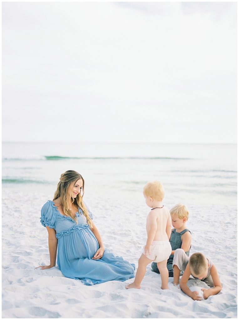 Pregnant mother sits on the beach looking at her three young sons who are playing in the sand