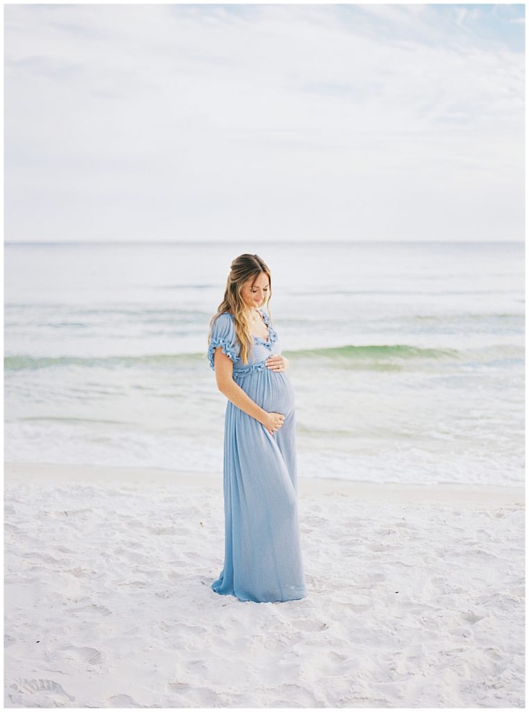 Mother stands on the beach in a blue dress with her hands below her belly during her beach maternity session