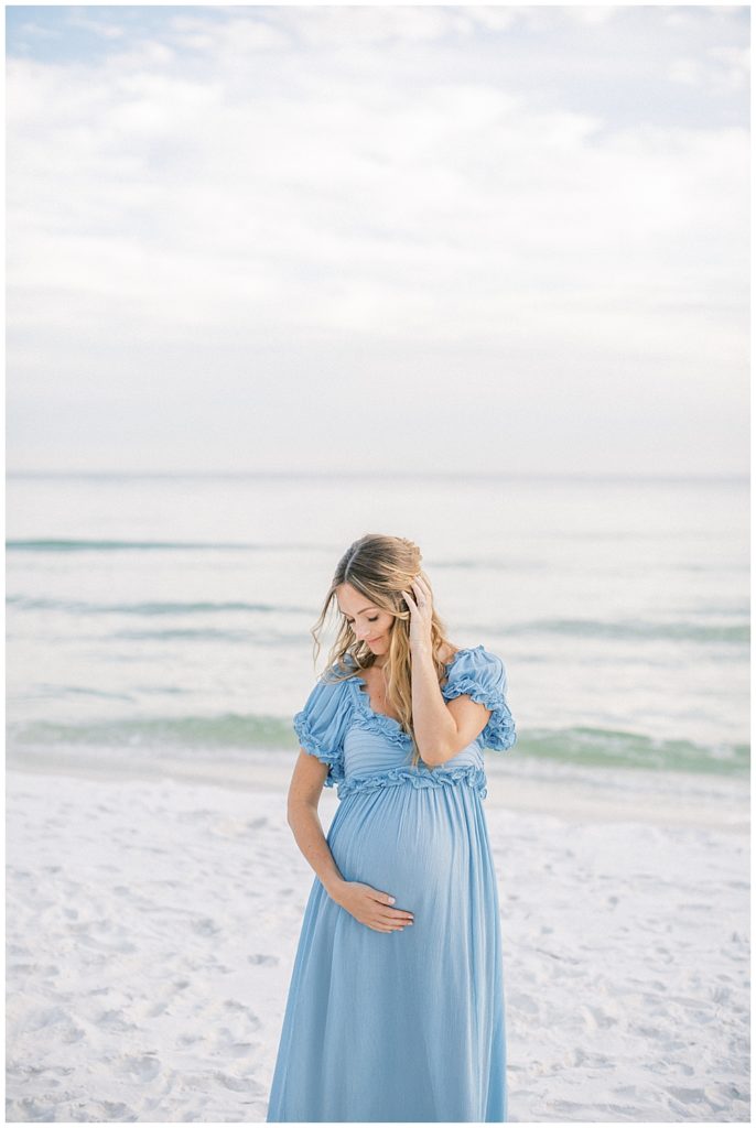 A pregnant mother stands on the beach with one hand in her hair and one hand below her belly during her beach maternity session