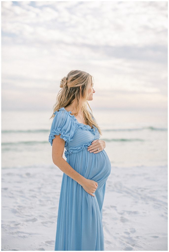 Pregnant woman in blue Doen dress stands on the beach looking out into the water during her beach maternity photoshoot