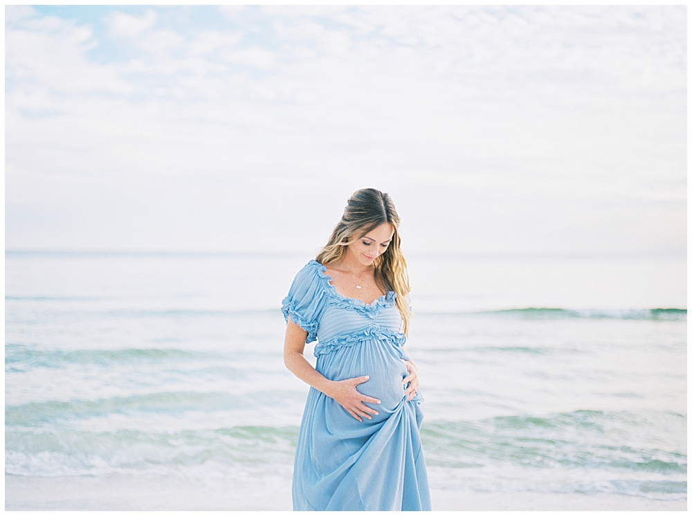A mother in a blue dress stands on the beach with both hands resting below her belly