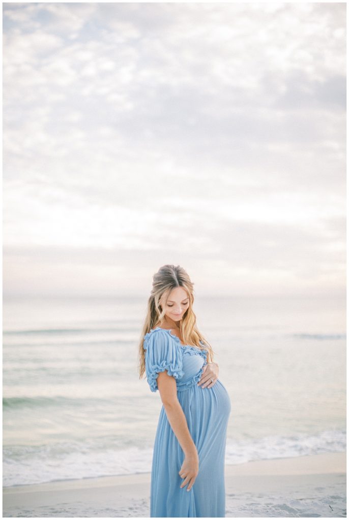 Pregnant mother stands with one hand on top of her belly and one hand holding her blue dress during her beach maternity session