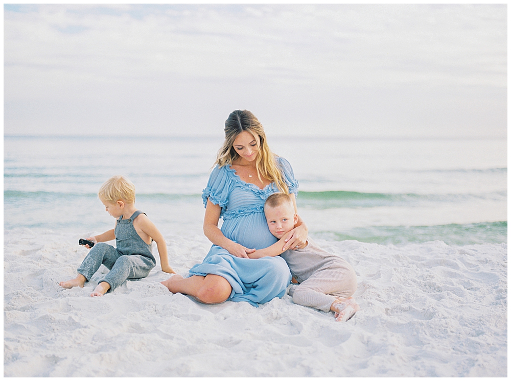 A mother in a blue dress sits on the beach during her maternity session with her two young boys, one of whom is hugging her