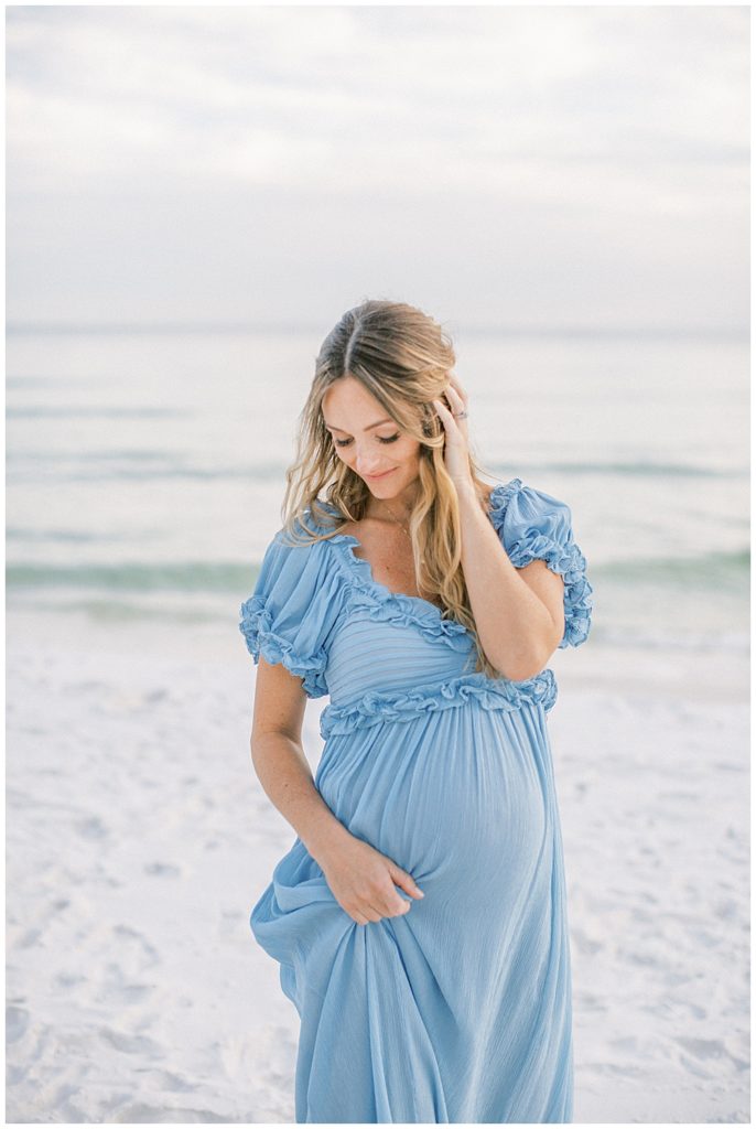 Pregnant mother stands on the beach with one hand tugging her dress and one hand in her hair during her beach maternity photoshoot