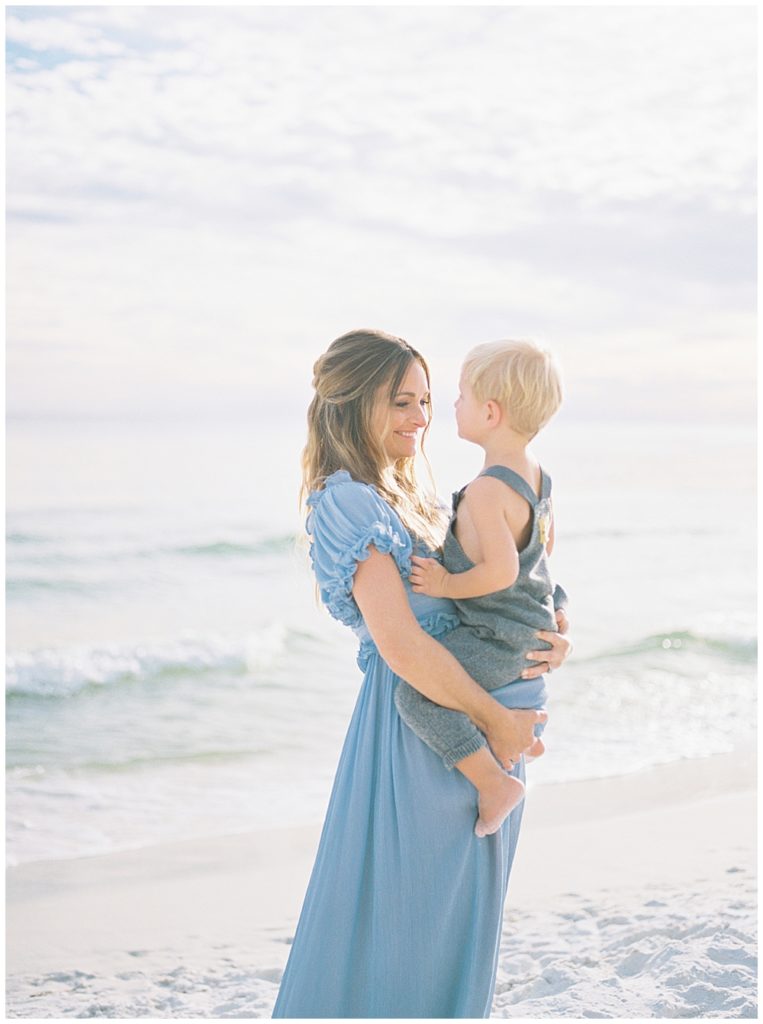 Mother holds her young son on the beach and smiles at him during her beach maternity photoshoot