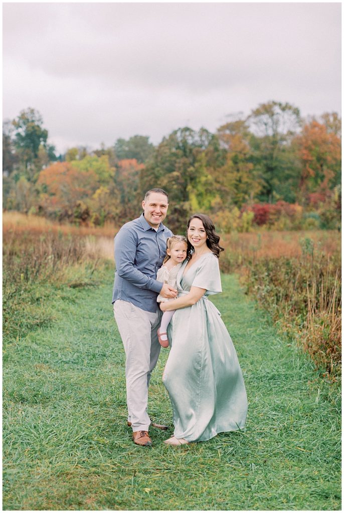 Mother and father hold their toddler daughter while standing in a clearing at the Howard County Conservancy during their fall family photos by Maryland Family Photographer Marie Elizabeth Photography