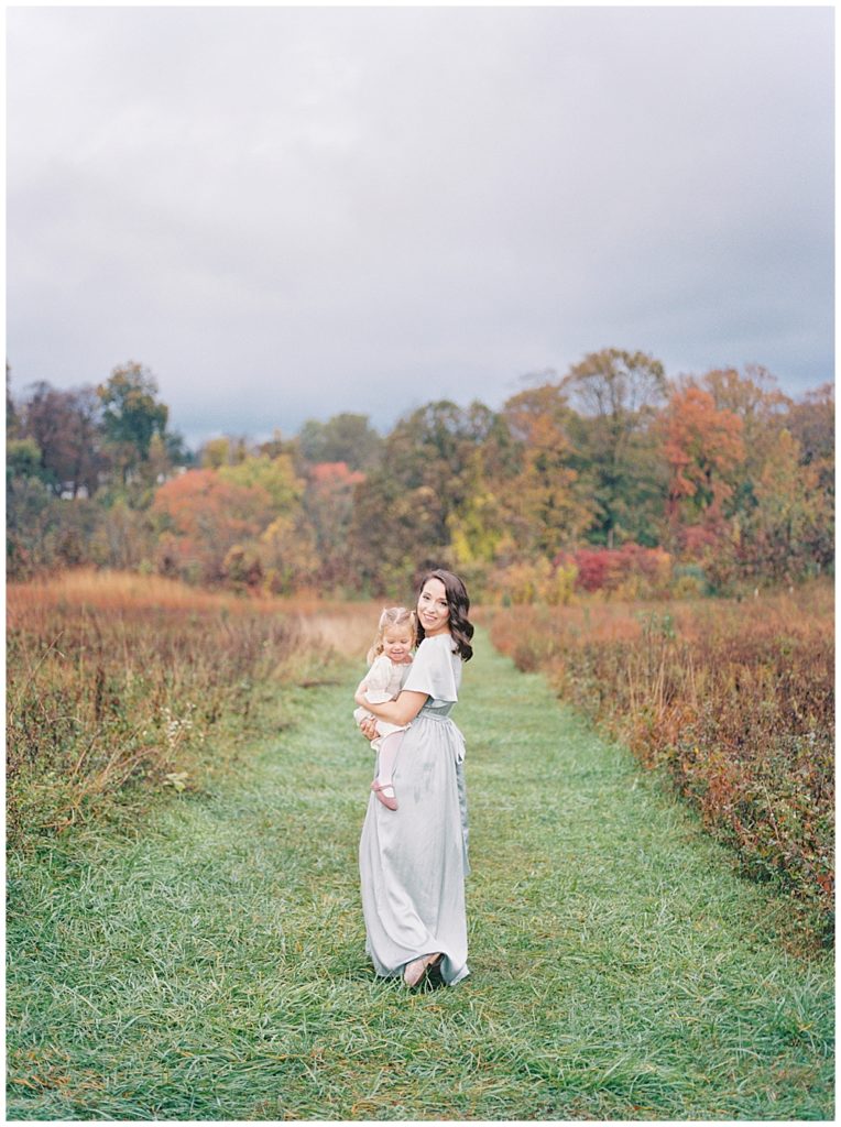 Mother walks holding her toddler daughter in the Howard County Conservancy during their fall session and turns to look back at the camera