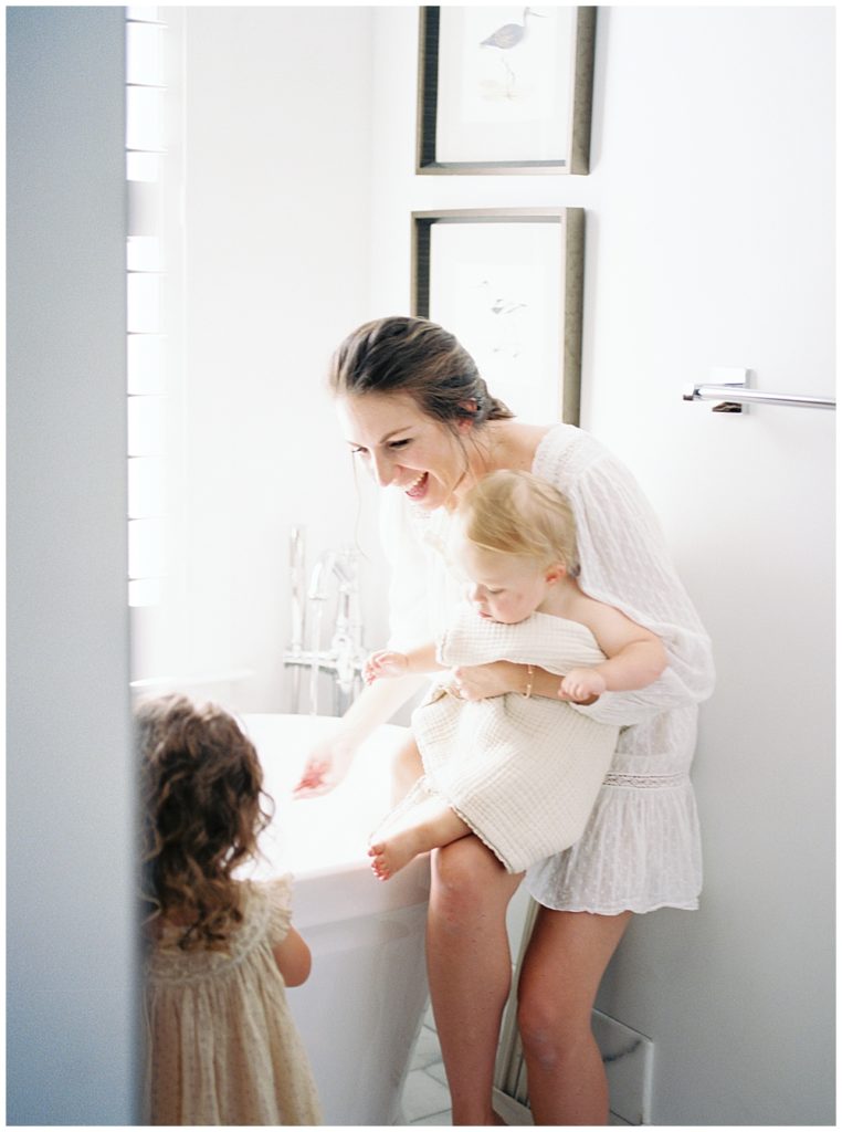 Mother sits on the edge of the tub while holding her baby and smiling at her toddler daughter standing in front of her