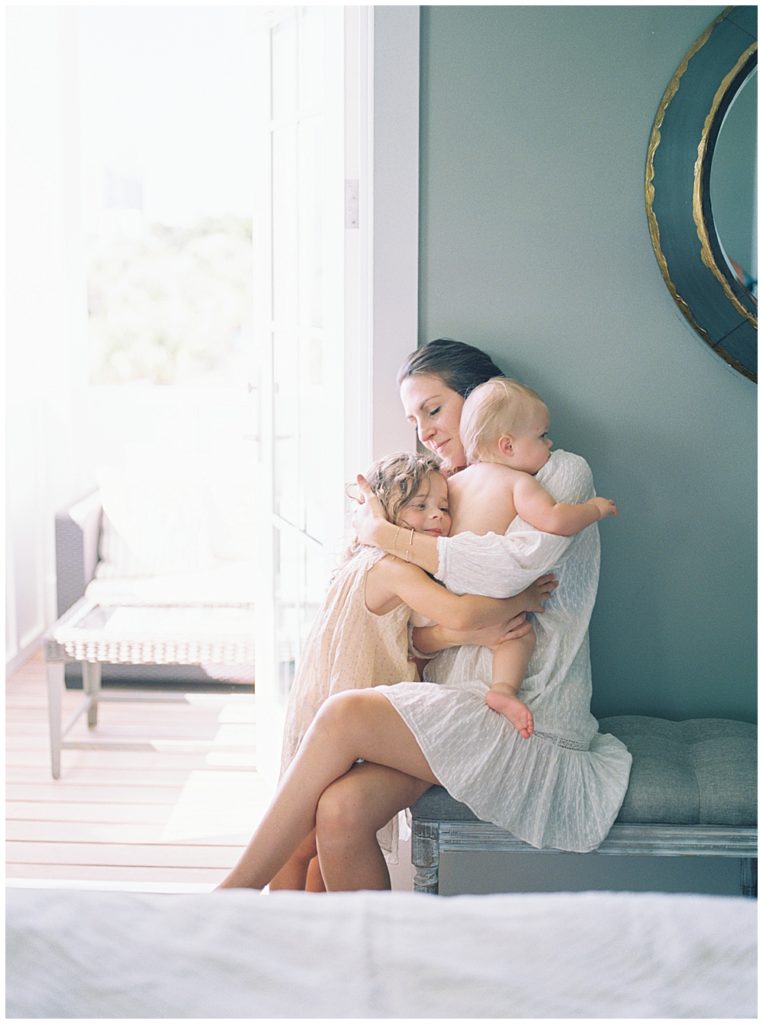A mother sits on a bench in her bedroom and hugs her toddler daughter and infant daughter