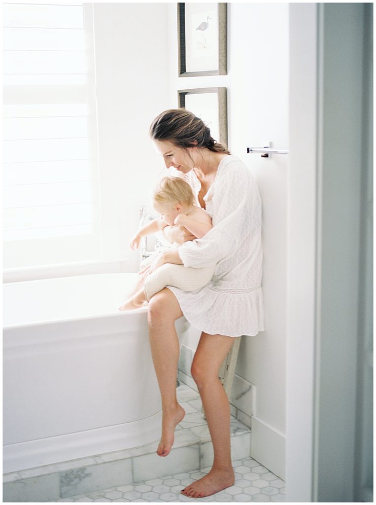A mother sits on the edge of the tub in the bathroom holding her infant daughter on her lap during her lifestyle family session at her home