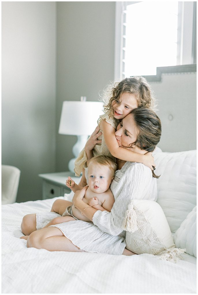A little girl stands on the bed an hugs her mother while her mother sits and holds her other daughter on her lap during their family photo session