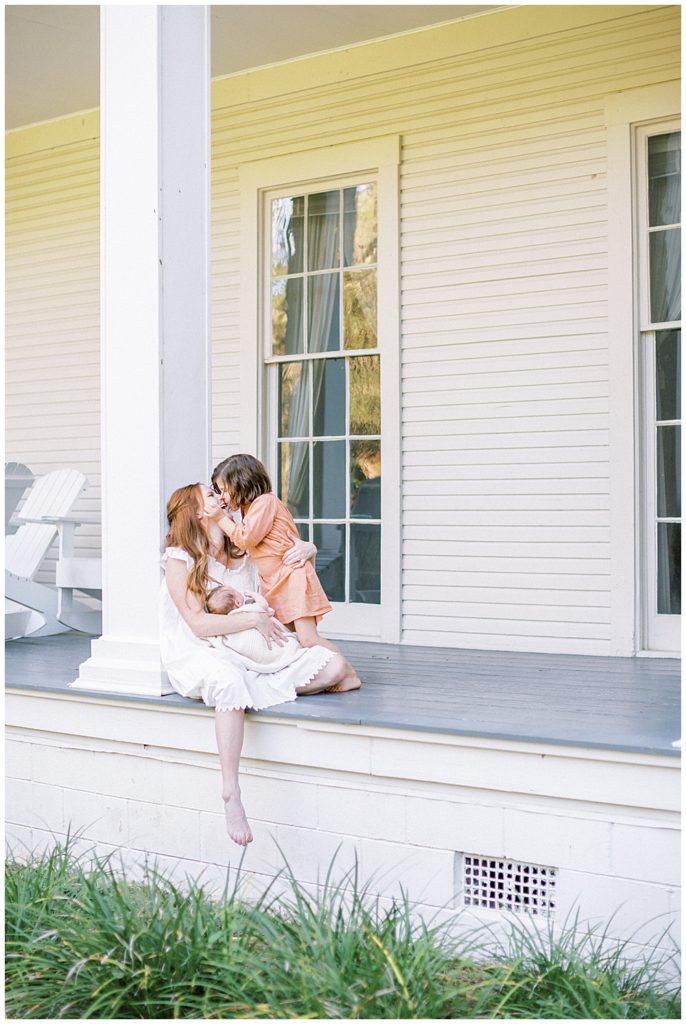 A little girl goes over and gives her mother, who is holding a baby, a kiss while sitting on the porch