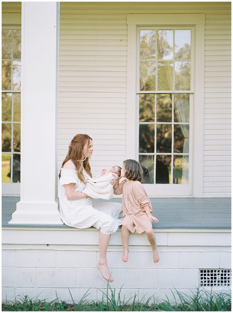 A mother sits on the front porch of a manor with her daughter who leans over and kisses the mother's newborn baby on the head
