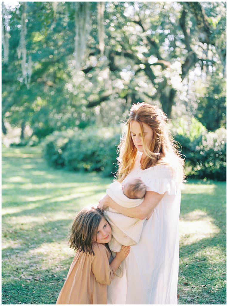 A red haired mother stands outside in a green park holding her newborn while her young daughter gives her a hug