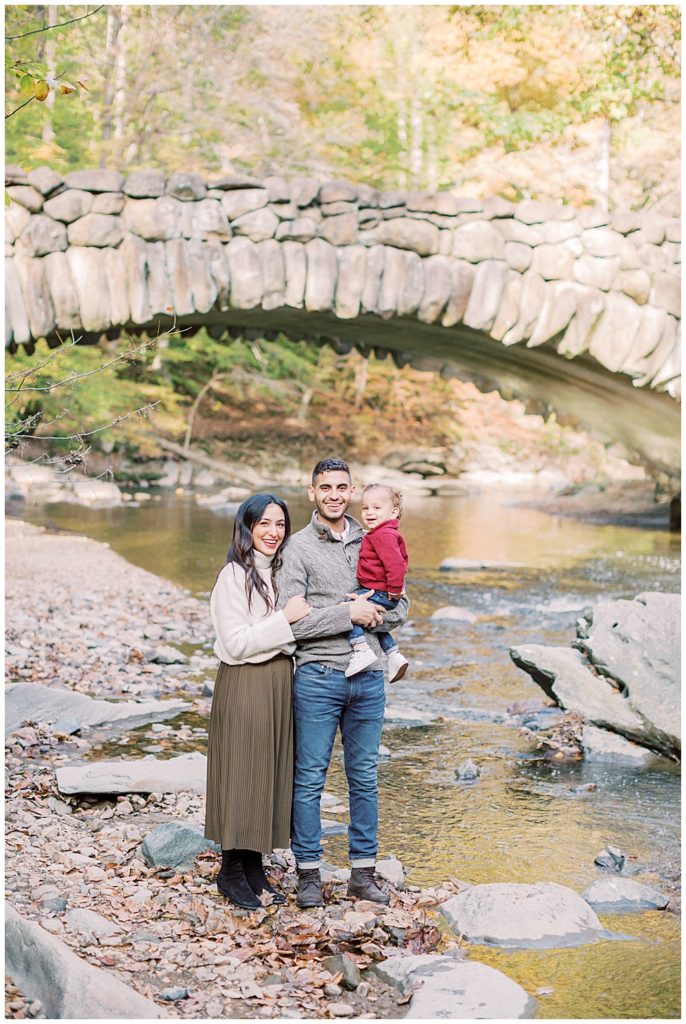 A mother, father, and baby boy smile at the camera while standing in front of Boulder Bridge in DC