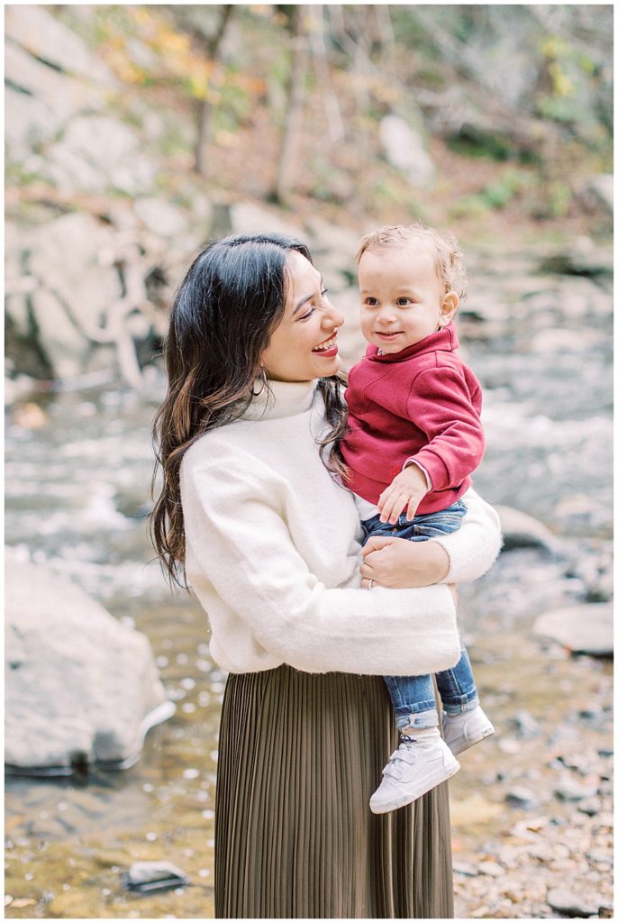 Mother smiles and holds her baby in a red sweater by a creek