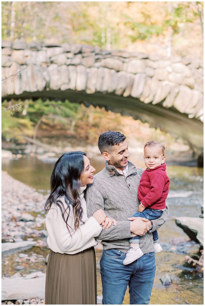 Mother and father hold their infant son in front of Boulder Bridge in Rock Creek Park while smiling at him