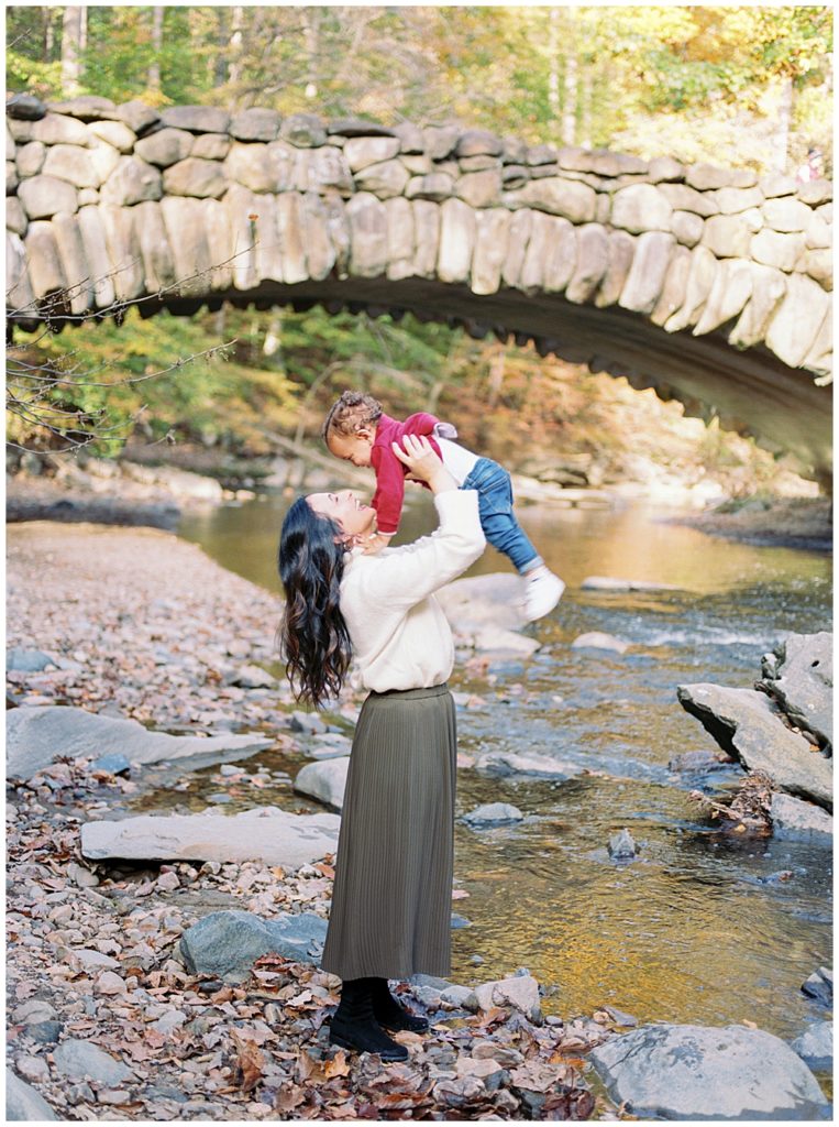 A mother holds up her baby boy in the air while standing on rocks in front of Boulder Bridge in Washington DC