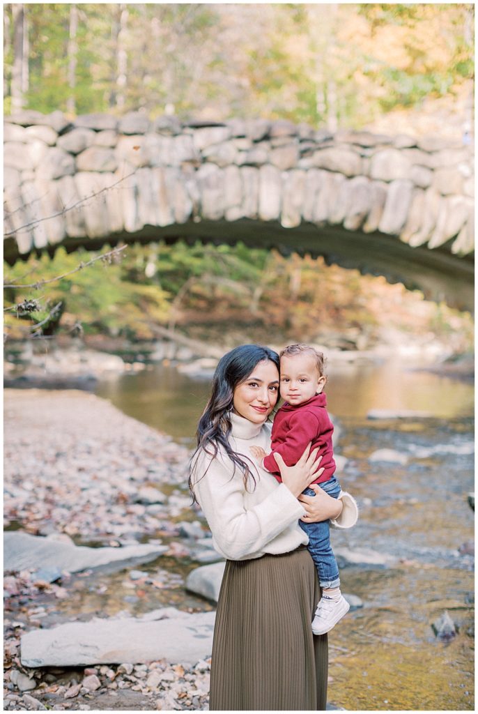Mother holds her son close to her standing in front of Boulder Bridge in Rock Creek Park in DC