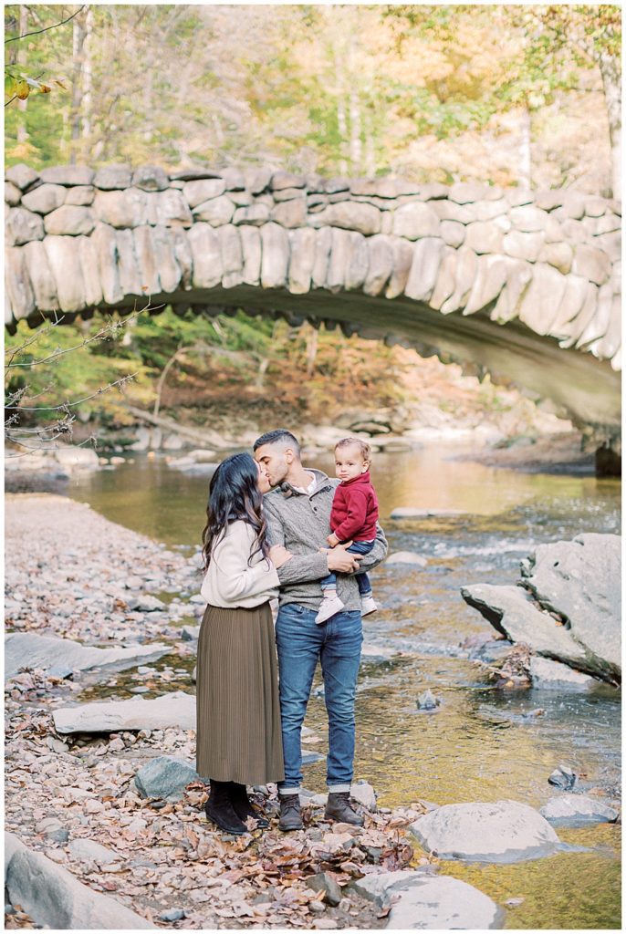 Mother and father kiss while holding their baby boy in front of Boulder Bridge in Rock Creek Park in DC