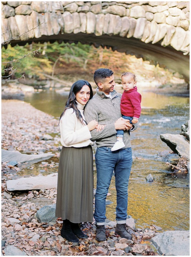 Mother, father, and baby boy stand in front of Boulder Bridge at Rock Creek Park in DC
