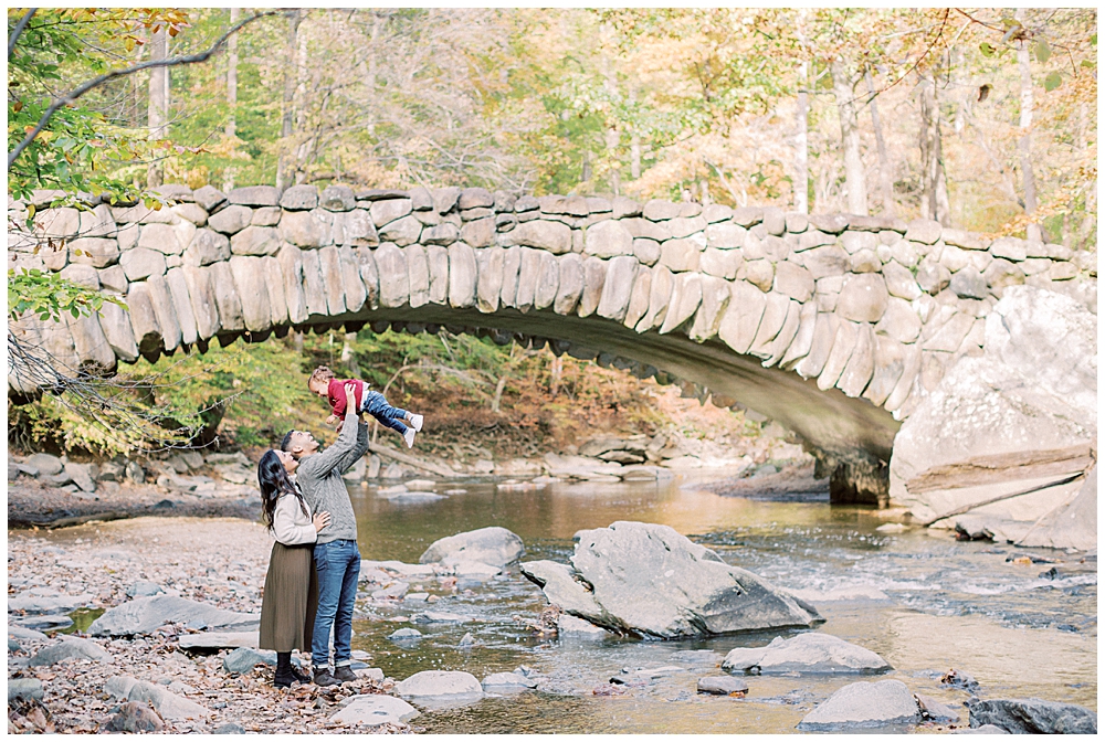 Mother and father hold their baby boy up into the air in front of Boulder Bridge in Rock Creek Park in DC