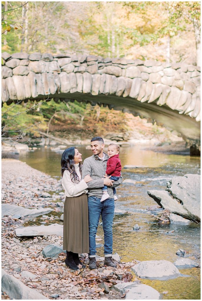 Mother and father look lovingly at one another while holding their one year old son in front of Boulder Bridge in Washington DC