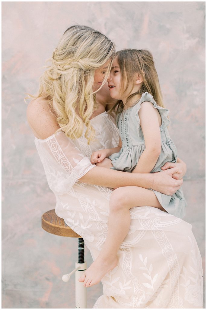 A mother in a white dress and her daughter in a green dress sit on a stool and nose nuzzle during their studio motherhood session in Bethesda Maryland