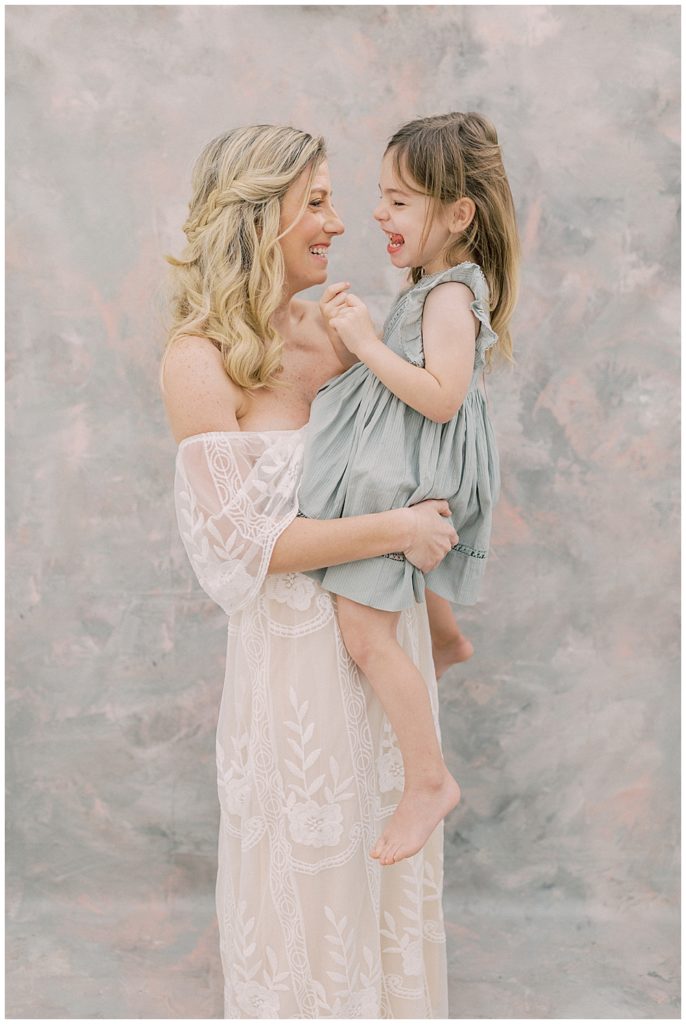 A blonde mother in a white dress holds her little girl in a green dress and smiles