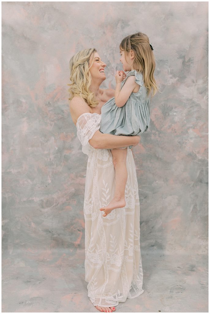 A blonde mother in a white dress holds her little girl while they both laugh