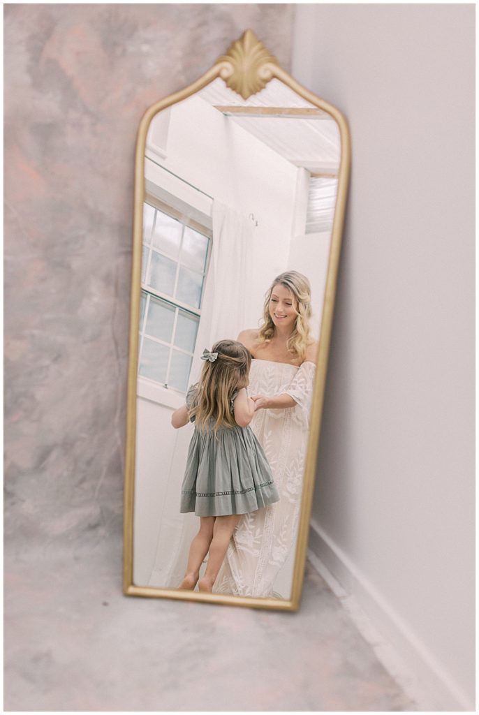 A mother dances with her daughter as seen through a gold trimmed mirror during their studio motherhood session