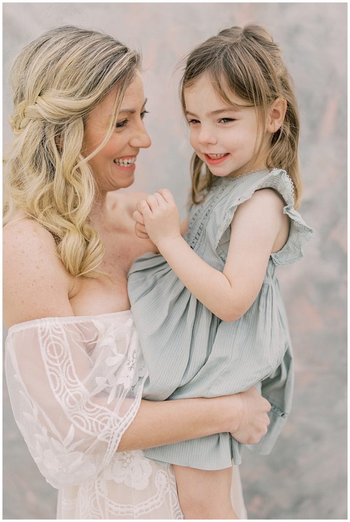 Blonde mother in a white dress smiles while holding her little girl in a green dress