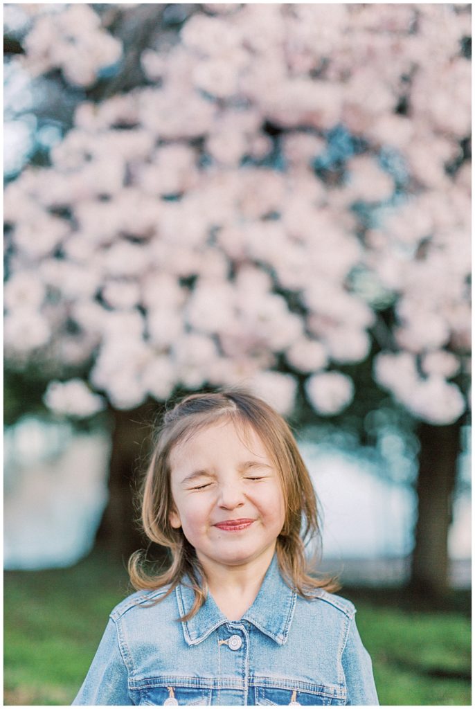 Little girl smiles and closes her eyes in front of a cherry blossom tree