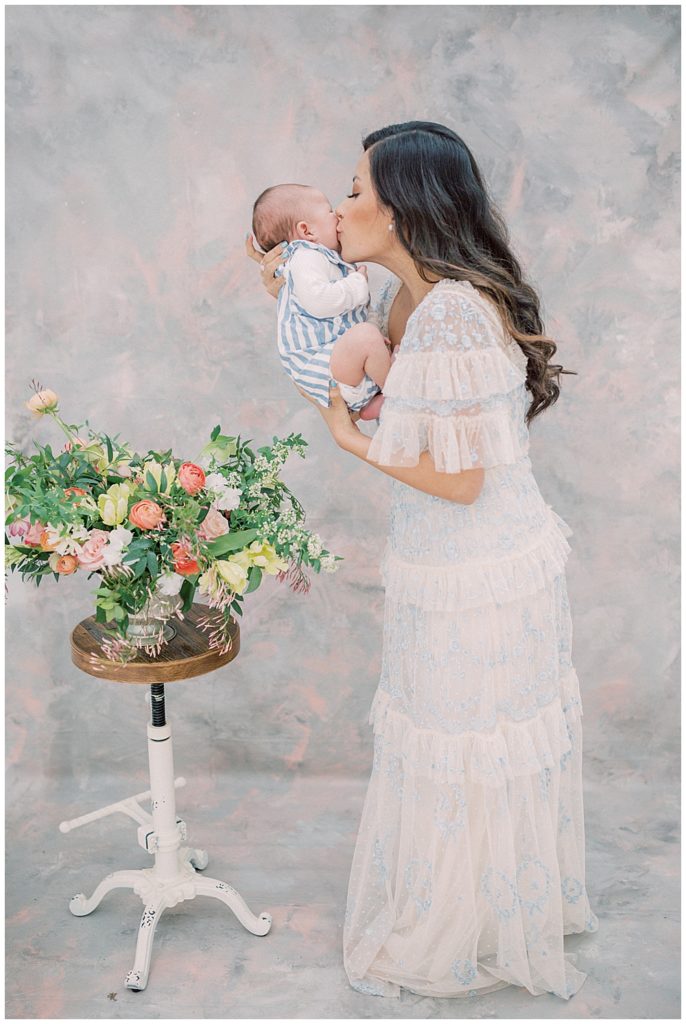 Mother kisses her baby boy next to a floral display during her motherhood studio session