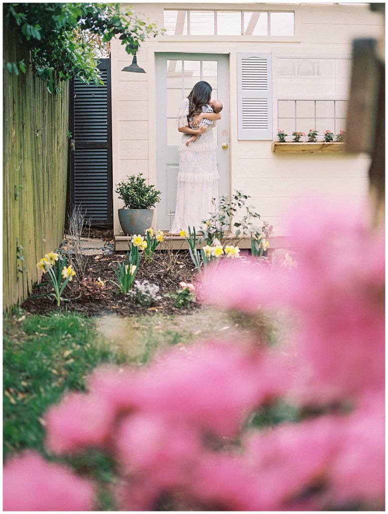 Mother kisses her infant son while standing in front of a blue door with pink flowers in the foreground