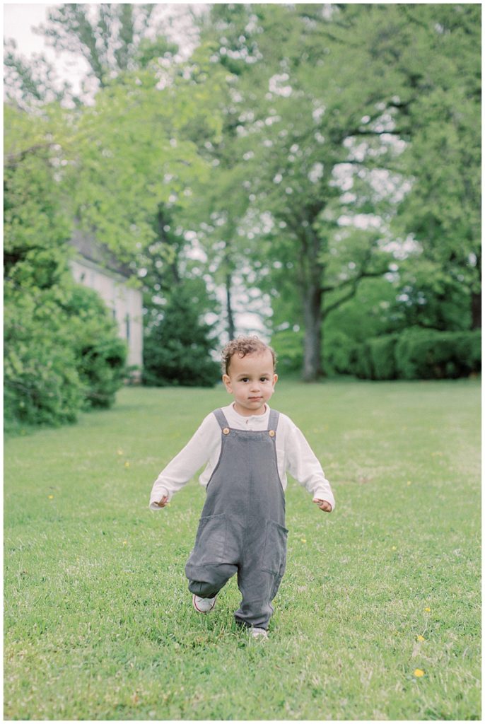 Toddler boy in overalls runs in the grass at Salubria